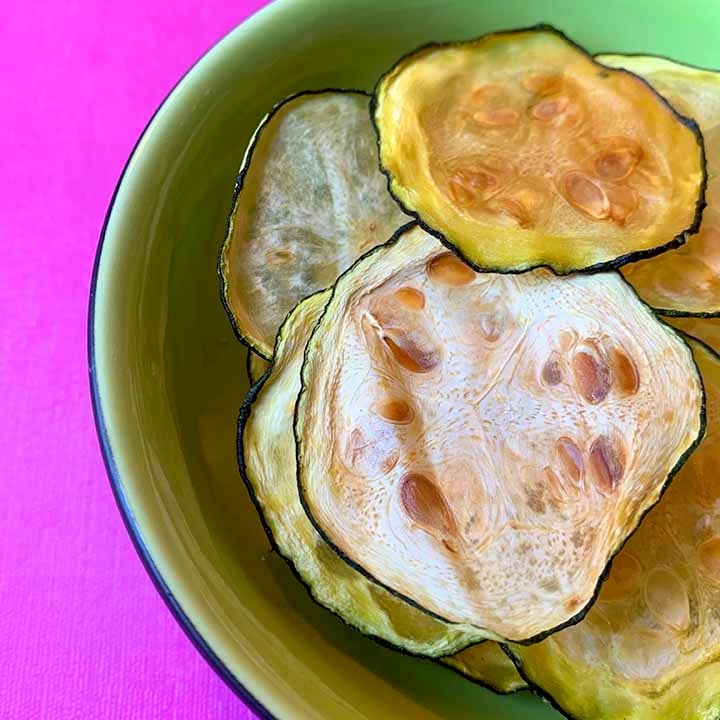 A reen bowl of low carb zucchini chips agaisnt a hot pink background