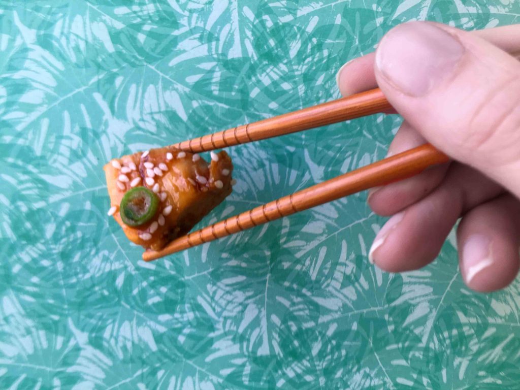 a hand holds a piece of Keto Sesame Chicken in a pair of chopsticks