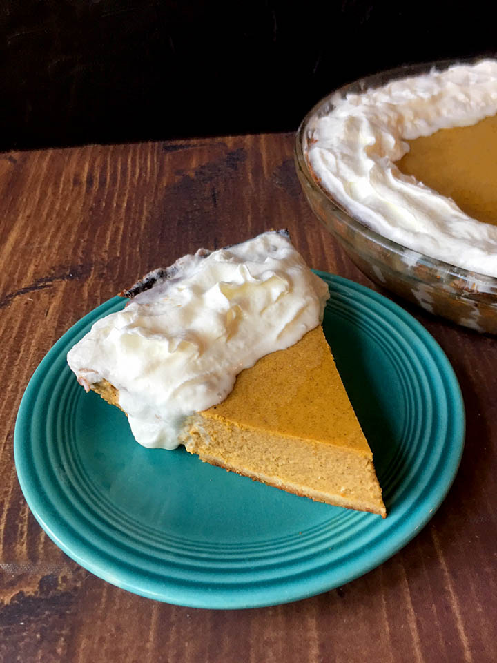 a slice of Keto pumpkin pie on a blue plate with a pumpkin pie in the background