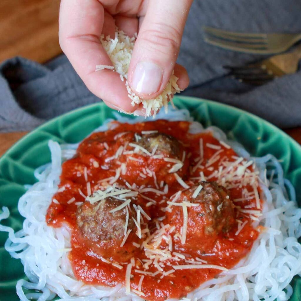 a hand sprinkles some cheese on Keto Spaghetti and Meatballs
