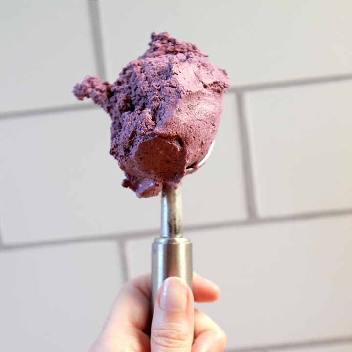 A hand holds an ice cream scoop of Sugar-Free Blueberry Ice Cream against a tile background