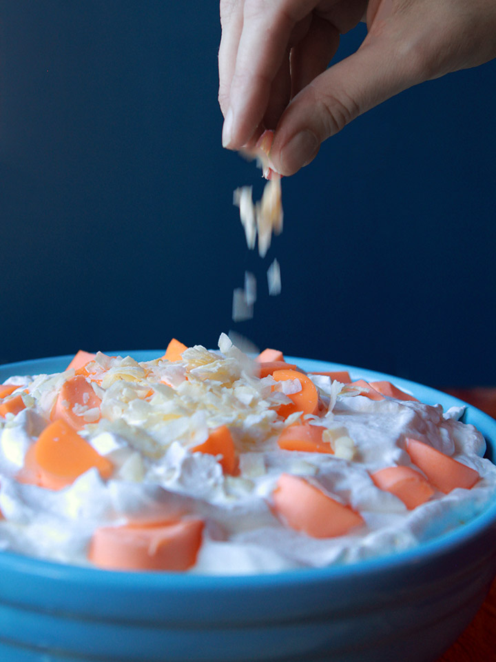 a hand sprinkles coconut flakes on a bowl of orange fluff salad