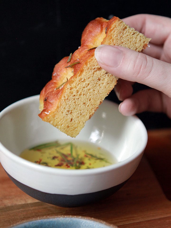 a hand dips a slice of low carb yeast bread focaccia into a bowl of herbed olive oil